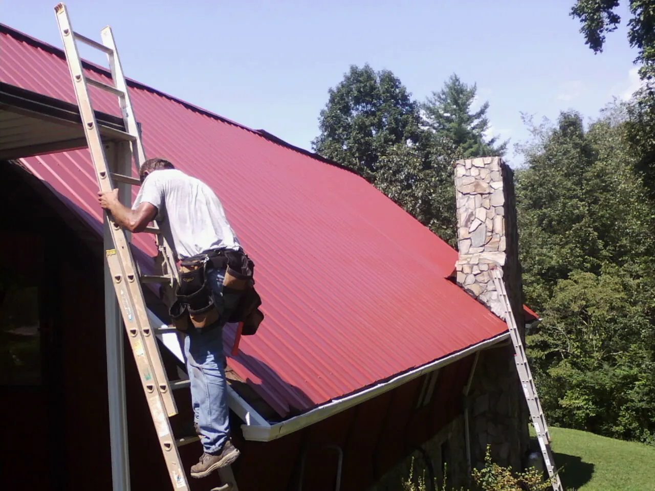 Roofer on a ladder beside a red metal roof and stone chimney — Soffit & Fascia Repair by Roofing Brothers in Parrott